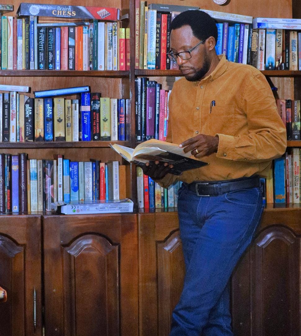 Valentino Kamya reading a book beside his bookshelf